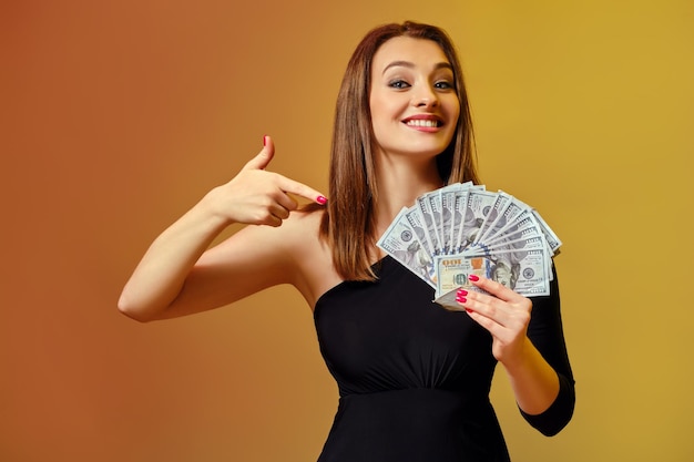 Prachtige blonde meisje met lichte make-up rode manicure in zwarte jurk is glimlachend en wijzend op fan van honderd-dollarbiljetten in haar handen poseren op kleurrijke achtergrond Poker casino close-up