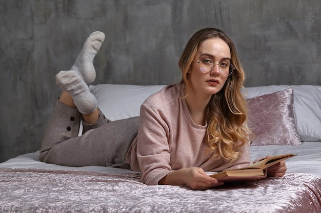 Prachtige blonde vrouw in glazen, casual kleding. Ze kijkt naar je, houdt een boek vast en ligt op bed. Student, blogger, document studeren. Interieur met grijze muur, roze deken en kussens. Detailopname