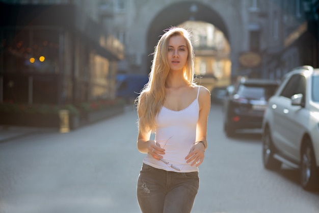 Prachtige blonde vrouw met lang haar, gekleed in jeans en t-shirt, wandelen in de stad in zonnestralen