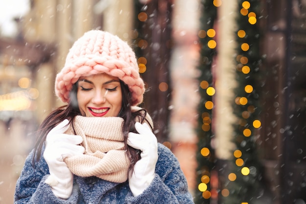 Prachtige brunette vrouw draagt gebreide lichtroze muts en sjaal wandelen in de stad tijdens de sneeuwval. Ruimte voor tekst