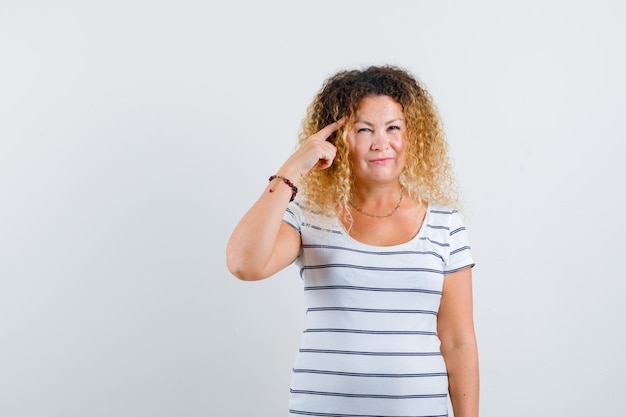 Prachtige dame die de vinger op de tempel houdt in een gestreept t-shirt en er zelfverzekerd uitziet. vooraanzicht.