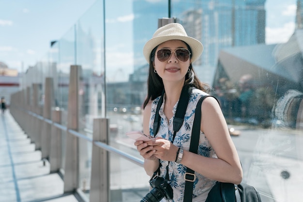 prachtige jonge vrouw met zonnebril messaging op slimme telefoon in stad straat achtergrond. meisje met mobiel gesprek in zonnevlam. reiziger draagt camera leunend op raam van viaduct in de stad