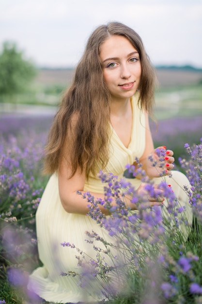 Prachtige jonge vrouw van 25 jaar met prachtig lang haar, poserend in een romantische gele zomerjurk op het lavendelveld