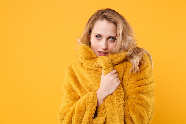 Prachtige mooie jonge blonde vrouw meisje poseren geïsoleerd op oranje muur achtergrond, studio portret. Mensen oprechte emoties levensstijl concept. Bespotten kopie ruimte. Ze wikkelt zichzelf in een gele bontjas.