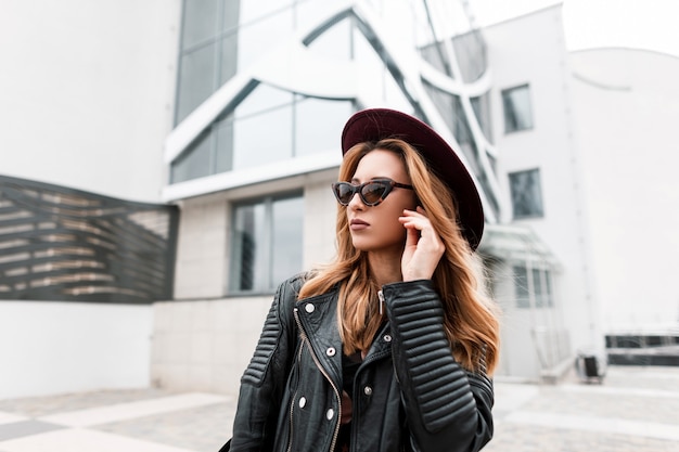 Prachtige roodharige hipster jonge vrouw in vintage zonnebril in een stijlvolle hoed in een modieuze zwart lederen jas poseren