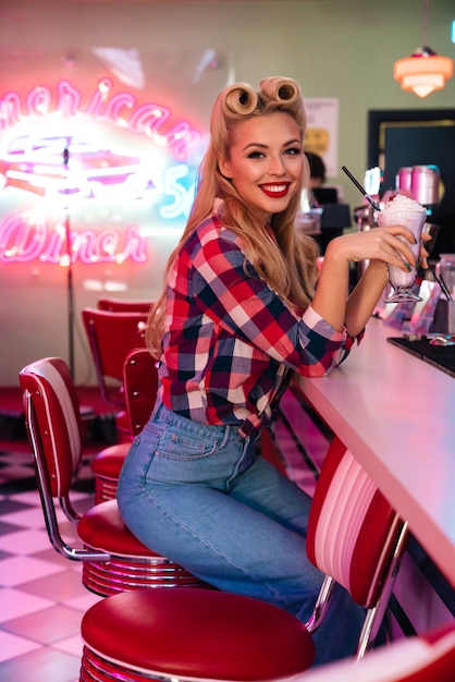 prachtige vrolijke vrouw met mooi kapsel die milkshake drinkt en lacht in retro amerikaans café