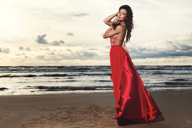 Prachtige vrouw, gekleed in mooie rode jurk op het strand tijdens zonsondergang tijd
