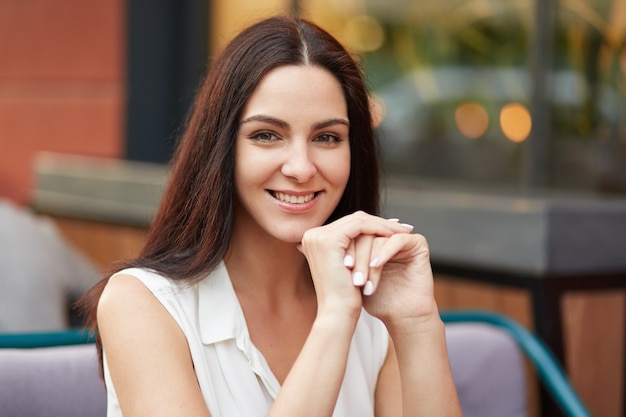 Prachtige vrouwelijke dame heeft een zachte glimlach, heeft donker haar, draagt een blouse, is in een goed humeur terwijl ze vrije tijd doorbrengt met vrienden bij de koffie