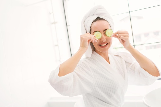 Prachtige zelfverzekerde volwassen vrouw met een handdoek op haar hoofd zitten en gesneden komkommers op haar ogen leggen in de badkamer thuis een mooie aantrekkelijke vrouw schoonheid levensstijl en cosmetisch concept