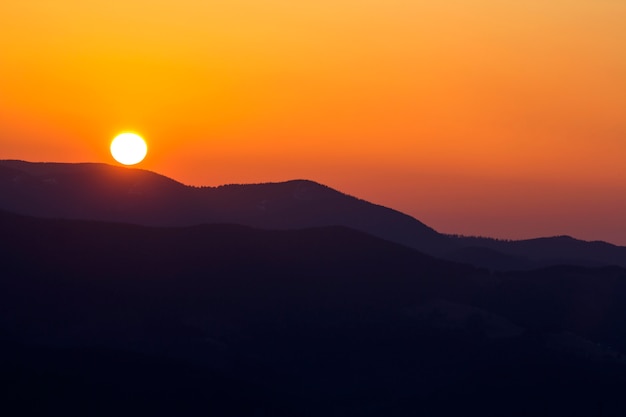 Prachtige zonsondergang in de bergen. Brede panoramamening van grote heldere witte zon in dramatische oranje hemel over donker bergketenlandschap bij zonsondergang of zonsopgang. Schoonheid van aardconcept.