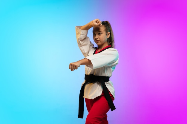 Practice. Karate, taekwondo girl with black belt isolated on gradient background in neon light. Little caucasian model, sport kid training in motion and action. Sport, movement, childhood concept.