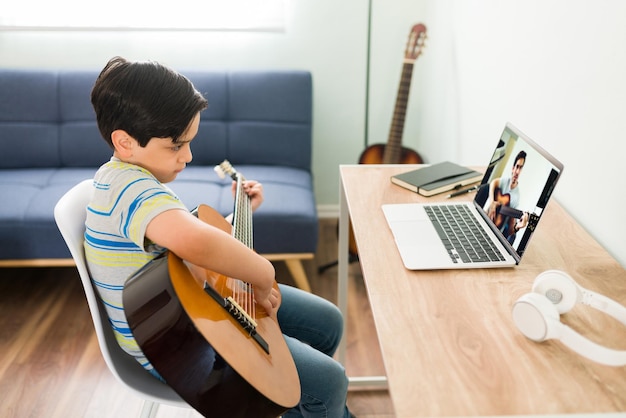 Practicing my acoustic guitar lessons. Caucasian kid sitting on his desk at home and practicing a new song on the guitar during his online music class