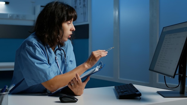 Practitioner assistant with blue uniform and stethoscope\
analyzing patient illness report working at health care treatment\
after hours in hospital office. nurse checking medical expertise on\
computer