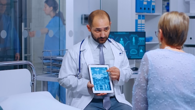 Practitioner doctor showing radiology scan on digital tablet to senior retired woman sitting on hospital bed in modern private clinic. Medical medicine explayining screen diagnosis scan treatment