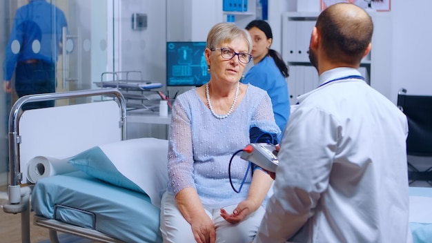 Il professionista misura la pressione sanguigna di una donna anziana in pensione con un monitor mentre l'infermiera lavora in background. sistema medico-sanitario, trattamento e diagnosi dell'esame della malattia