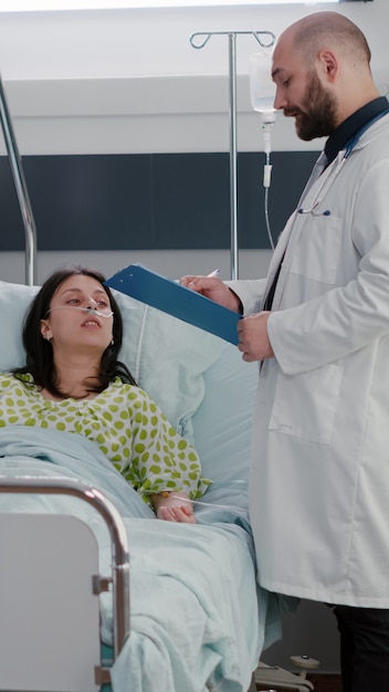 Practitioners doctors monitoring sick woman writing sickness treatment on clipboard during medical appointment in hospital ward. Patient resting in bed with nasal oygen tube explaining disease symptom