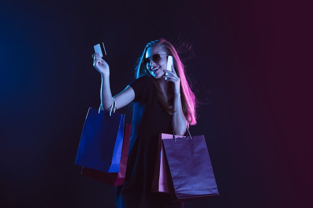 Pratende telefoon met boodschappentassen en kaart. Portret van een jonge vrouw in neonlicht op donkere achtergrondkleur. De menselijke emoties, zwarte vrijdag, cybermaandag, aankopen, verkoop, financieel concept.