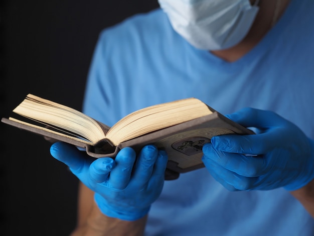 A prayer book in men's hands in blue medical gloves and a protective mask.