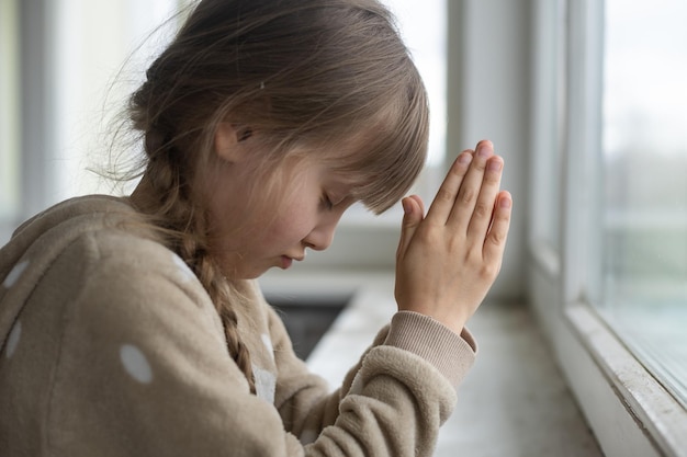 Bambina in preghiera vicino alla finestra