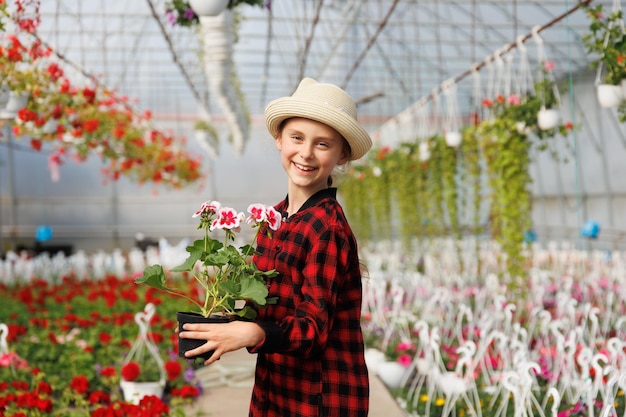 Pre-tiener meisje gelukkig meisje met hoed en een bloempot in haar hand glimlachend kind dat naar de camera kijkt is in de kas met veel bloemen Kopieer ruimte
