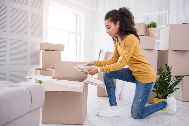 Precious memories. Charming curly-haired girl putting several framed photos into a box while packing her belongings before moving out