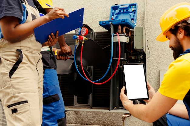 Precise serviceman cleaning and lubricating outdoor hvac system internal expansion valve, checking refrigerant level and calibrating thermostats while coworkers check maintenance steps on smartphone