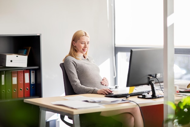 pregnancy, business, work and technology concept - smiling pregnant businesswoman with computer at office table