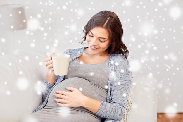 pregnancy, drinks, people, winter and expectation concept - happy pregnant woman with cup drinking tea at home over snow