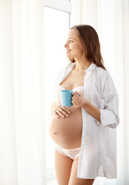 Gravidanza, bevande, riposo, persone e concetto di aspettativa - donna incinta felice in camicia e biancheria intima con tazza di tè che beve a casa