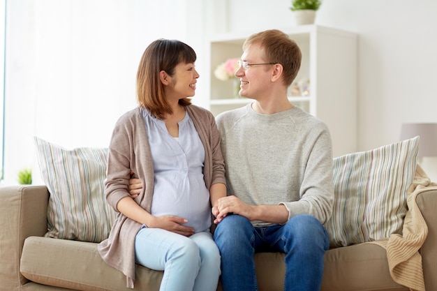 妊娠 家族と人々のコンセプト 幸せな妊娠した妻と夫が家でソファに座っている