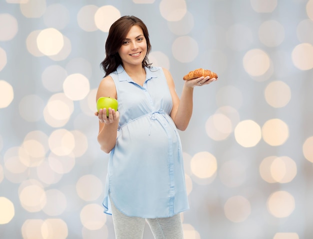 pregnancy, healthy eating, junk food and people concept - happy pregnant woman choosing between green apple and croissant over holidays lights background