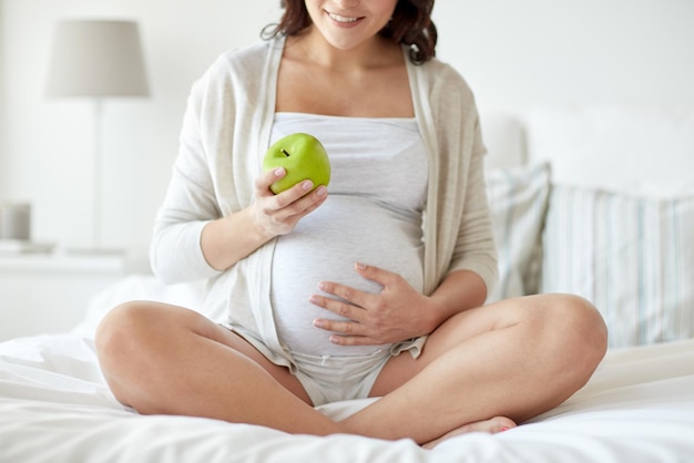 беременность, здоровое питание и концепция людей - крупный план счастливой беременной женщины, которая ест зеленое яблоко дома