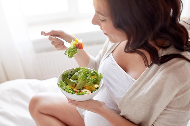 妊娠、健康食品、人々のコンセプト – 自宅のベッドで朝食に野菜サラダを食べる幸せな妊婦の接写