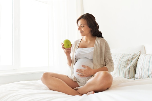 pregnancy, healthy food and people concept - happy pregnant woman eating green apple at home