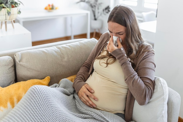 Pregnancy and illness. Sick pregnant woman blowing nose in tissue having fever sitting on sofa indoor suffering from covid-19