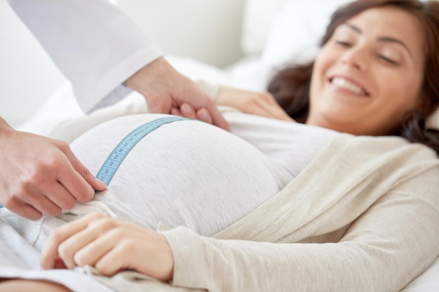 Photo pregnancy, medicine, health care and people concept - close up of obstetrician doctor with centimeter tape measuring pregnant woman tummy at hospital