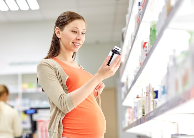 pregnancy, medicine, pharmaceutics, health care and people concept - happy pregnant woman reading label on medication jar at pharmacy