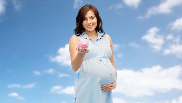 임신, 모성, 금융, 절약 및 사람 개념 - 푸른 하늘 배경 위에 돼지 저금통을 들고 있는 행복한 임산부