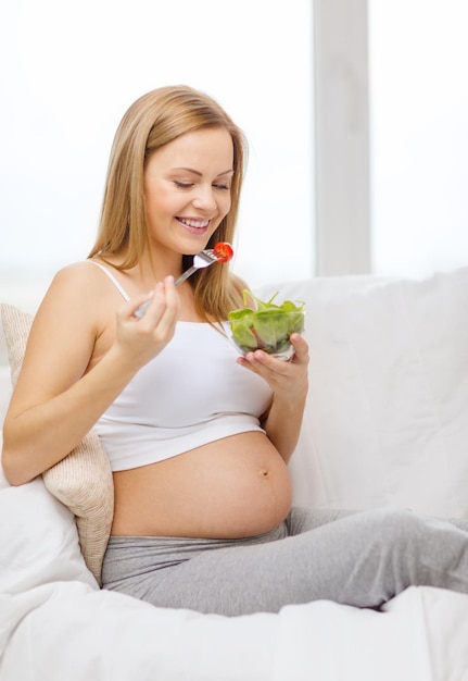 Photo pregnancy, motherhood, healthcare, food and happiness concept - happy pregnant woman sitting on sofa eating salad