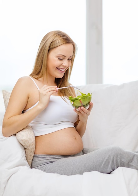 pregnancy, motherhood, healthcare, food and happiness concept - happy pregnant woman sitting on sofa eating salad