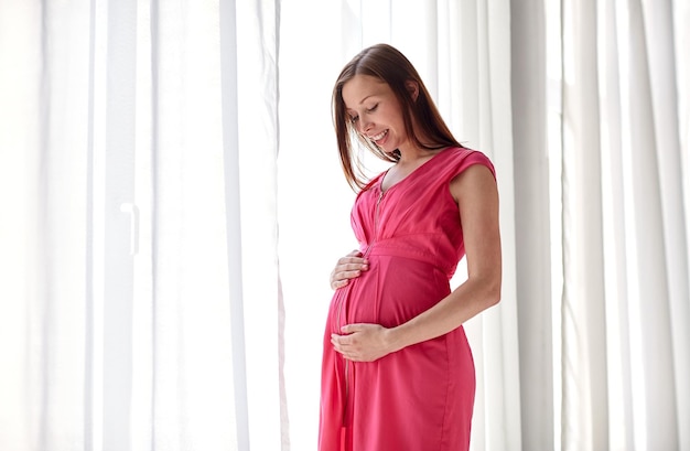 妊娠、母性、人々、期待のコンセプト – 家の窓から大きなお腹を持つ幸せな妊婦