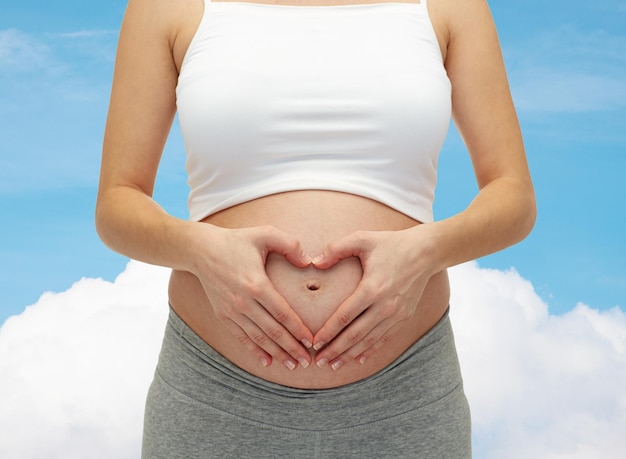 pregnancy, motherhood, people, love and expectation concept - close up of happy pregnant woman making heart shape hand sign on her bare tummy over blue sky and cloud background