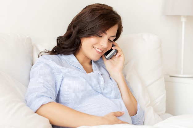 妊娠、母性、技術、人、期待のコンセプト – 自宅のベッドでスマートフォンを呼び出す幸せな妊婦