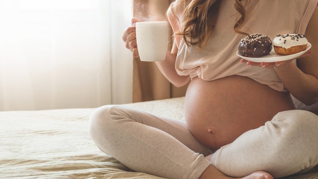 Беременность и питание. Беременная женщина, наслаждаясь пончиками и чаем в постели, свободное место