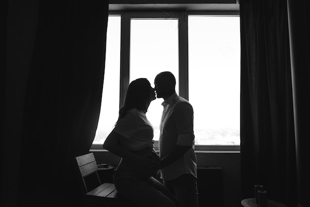 Pregnancy and people concept - happy man hugging his pregnant wife standing at window at home