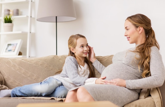 pregnancy, people and family concept - happy pregnant woman with girl sitting on sofa and talking at home