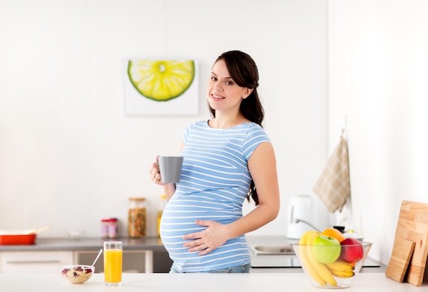 pregnancy, people and healthy eating concept - happy pregnant woman with cup of tea and food having breakfast at home kitchen