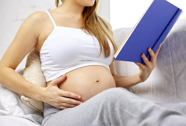 pregnancy, people and motherhood concept - close up of pregnant woman sitting on sofa and reading book