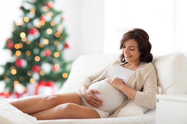 pregnancy, rest, people and expectation concept - happy pregnant woman lying on bed and touching her belly over christmas tree background