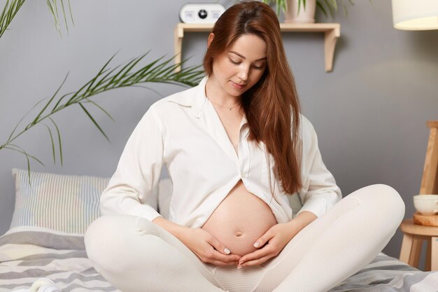 妊娠中の休息の人々と期待の妊婦が白い服を着て裸の腹を持ち、笑顔でベッドに座り、自宅で彼女の腹に触れる
